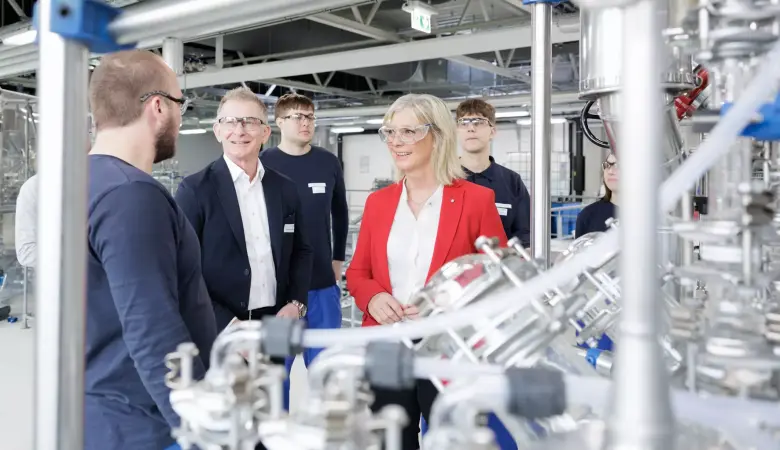 Die bayerische Arbeitsministerin steht in einem Kreis aus 4 Leuten und lächelt. Im Vordergrund ist eine große Aparatur aus Metall zu sehen.